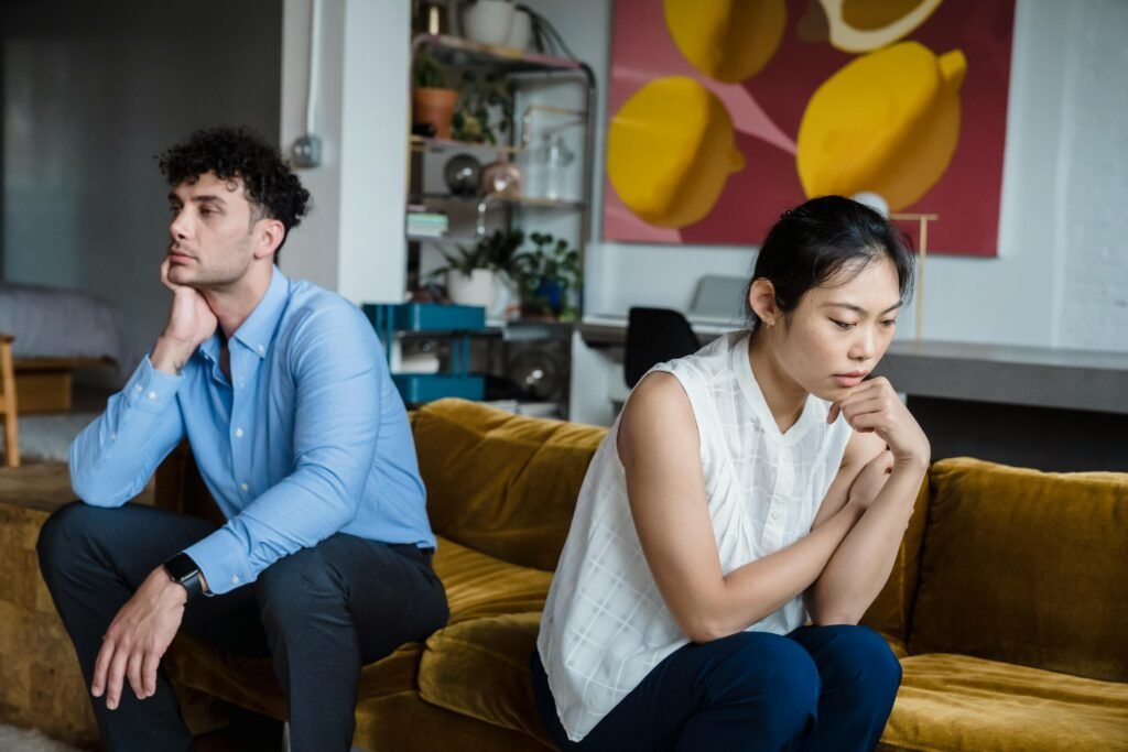 A couple sits indoors on a sofa, both appearing thoughtful and contemplative.